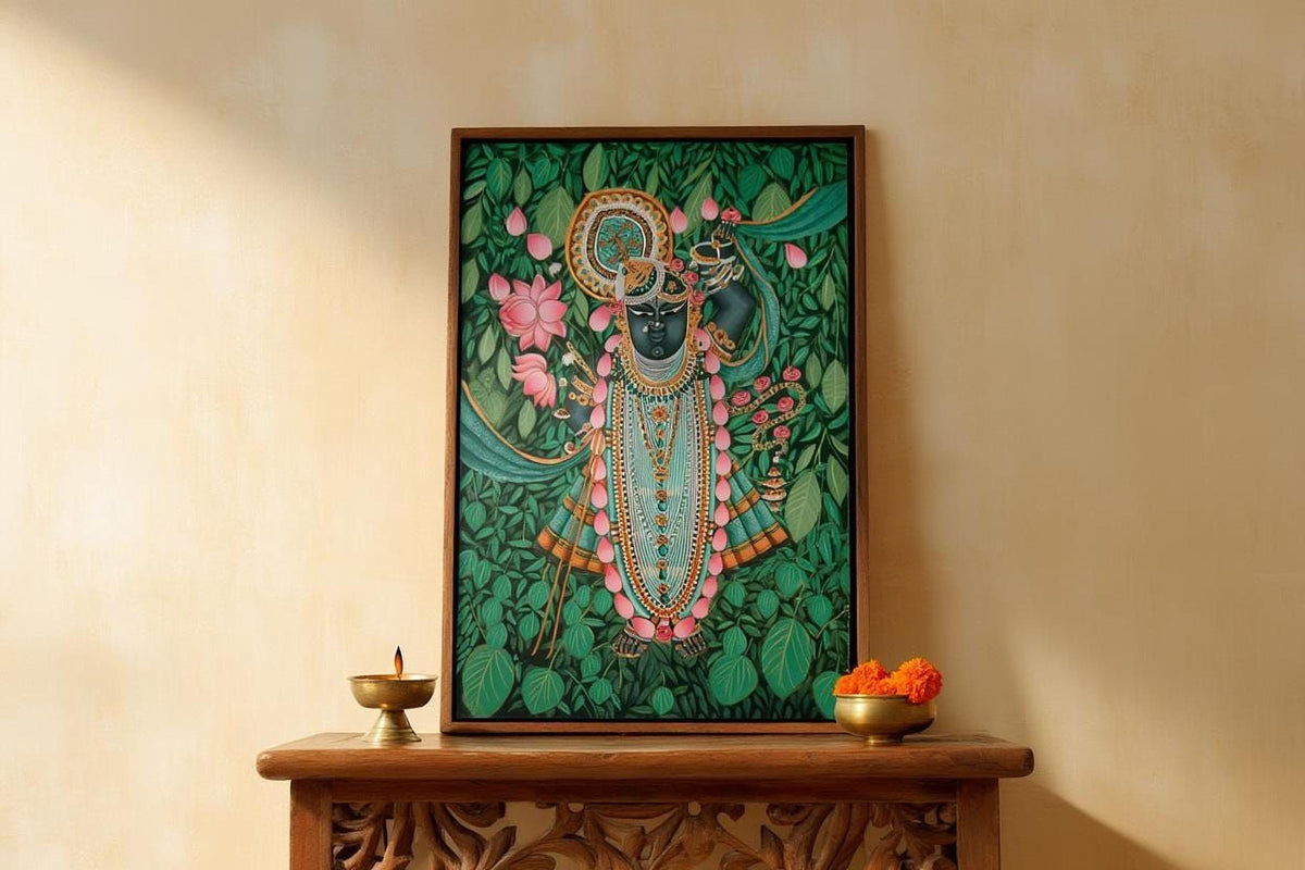a framed painting of a Hindu deity, surrounded by vibrant green leaves and flowers, placed on a wooden shelf. Below the painting, there are two small bowls containing orange flowers, and a small brass lamp.