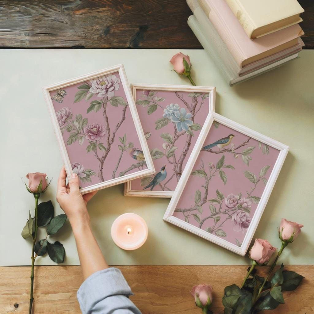 a person's hand holding a framed picture of a pink floral design, surrounded by a bouquet of pink roses, a lit candle, and a stack of books.