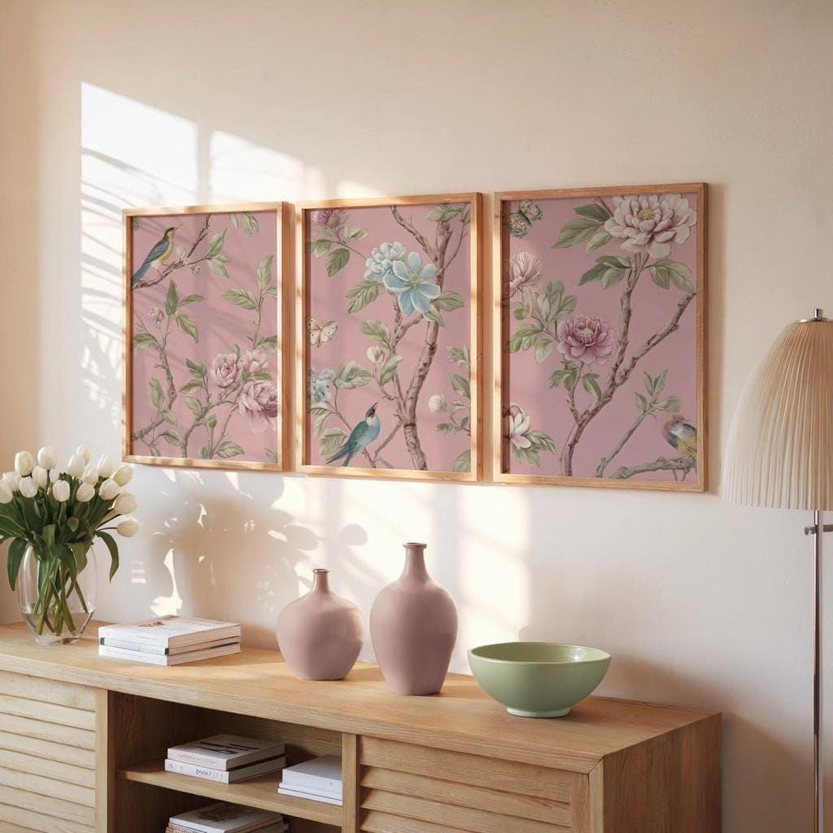 a wooden cabinet with three framed floral paintings hanging on the wall above it. The paintings depict various flowers and birds, creating a harmonious and visually appealing display. The cabinet also contains a vase, a bowl, and some books, adding to the overall aesthetic of the room.