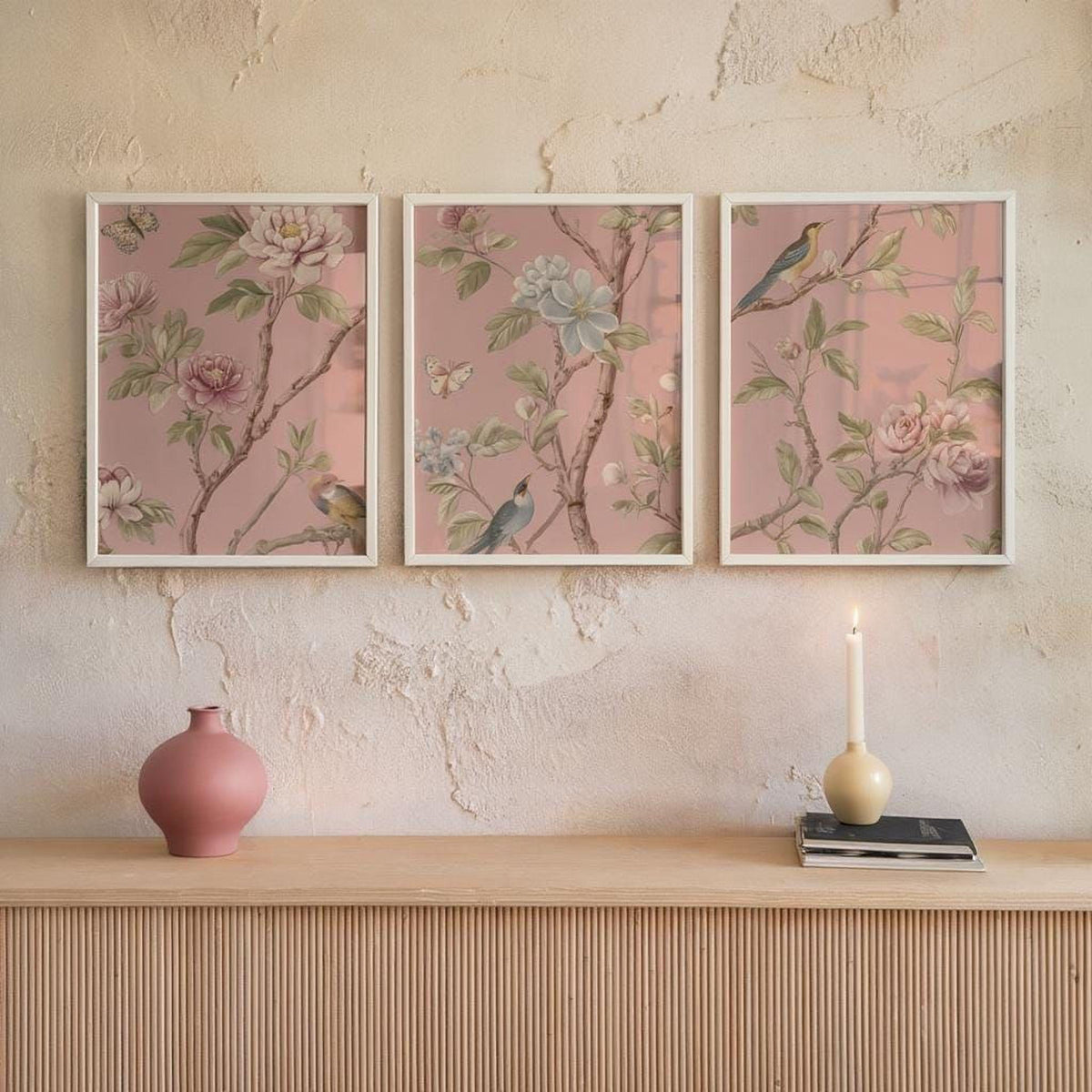 a shelf with three framed floral paintings hanging on the wall above it. On the shelf, there is a pink vase, a candle, and a stack of books.