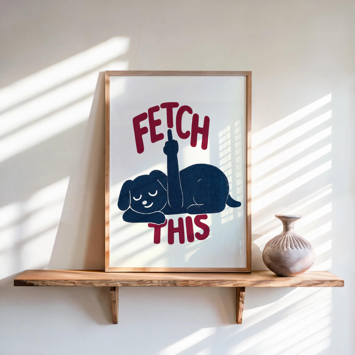 Framed print on a sunlit wooden shelf, dark navy dog and rust-red FETCH THIS text, retro dog artwork, funny dog wall art