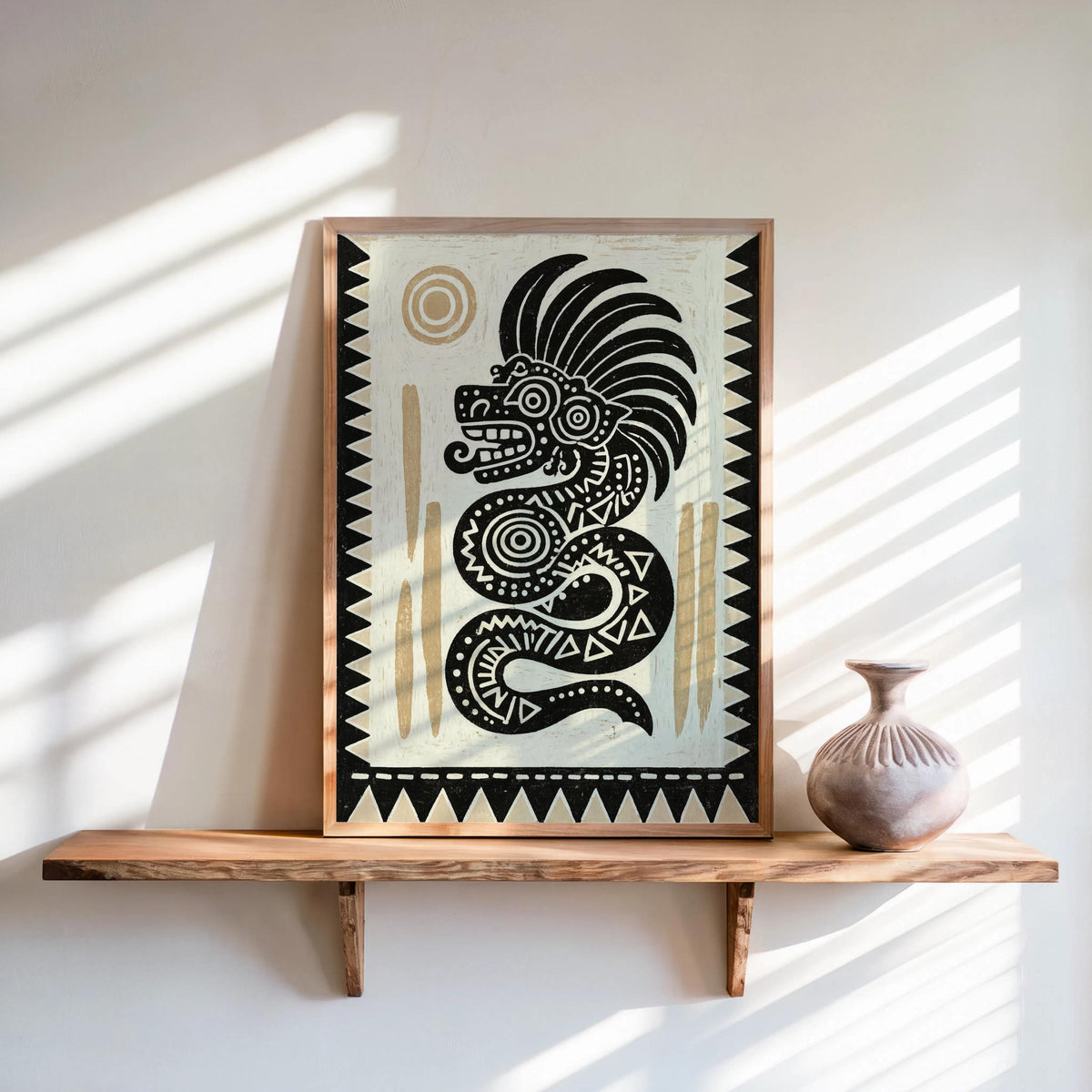 Framed Quetzalcoatl wall art leaning on a wood shelf, bold black-and-cream serpent with gold sun.