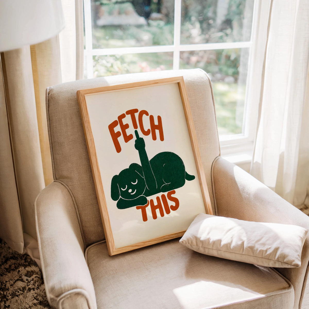 Framed print tucked into a beige armchair by a window, green dog silhouette with orange FETCH THIS lettering, cheeky dog print