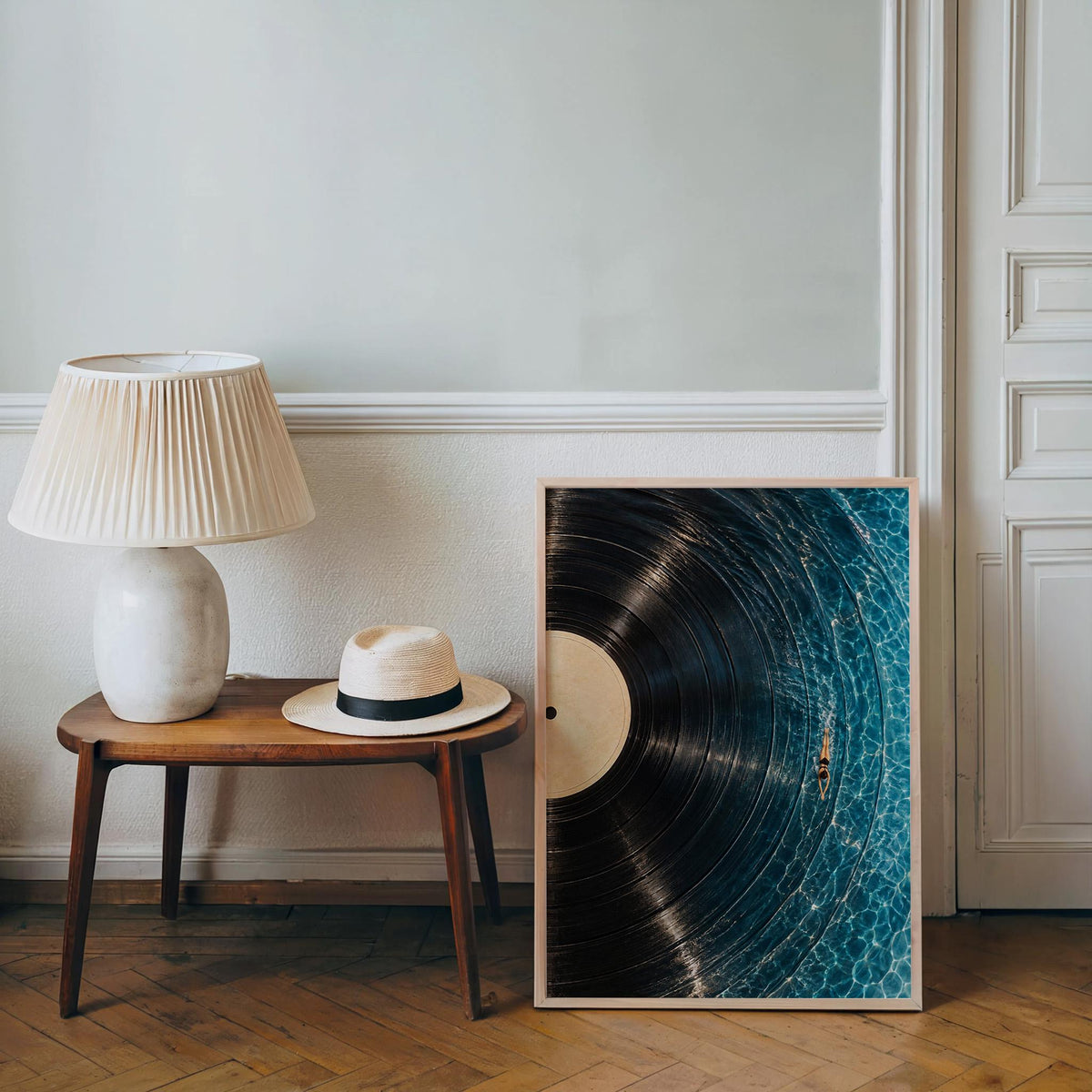 Framed vinyl record wall art propped by a side table and lamp, woven hat nearby, swimmer art print mood