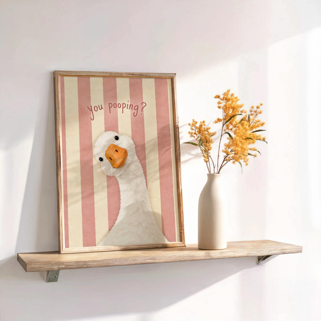 Framed pink striped funny goose bathroom print on shelf, whimsical pooping goose wall art styled with vase